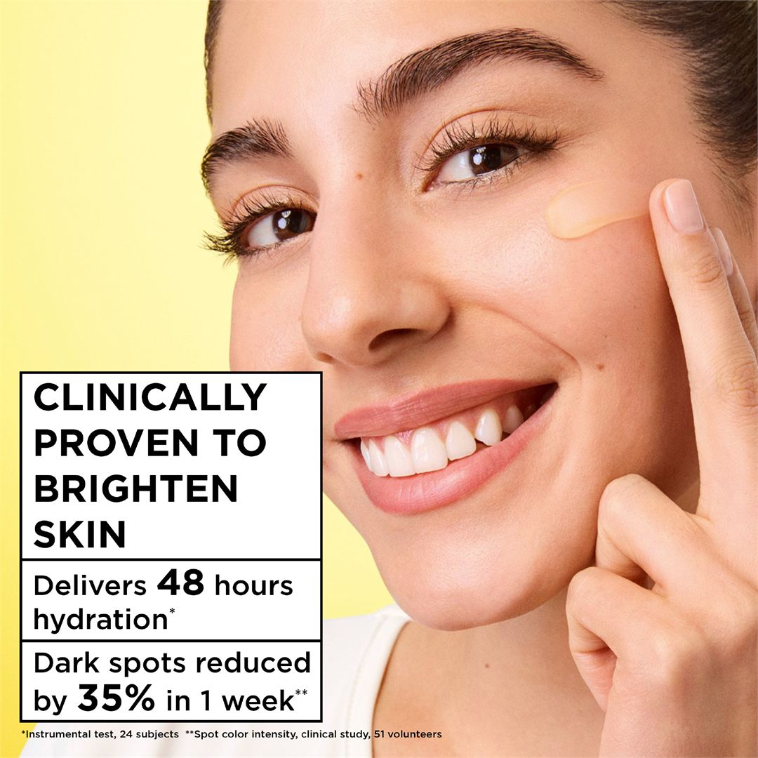 Close-up of a smiling woman applying or touching a beige skincare cream on her cheek, with text on the left reading 'Clinically Proven to Brighten Skin' and additional benefit claims.