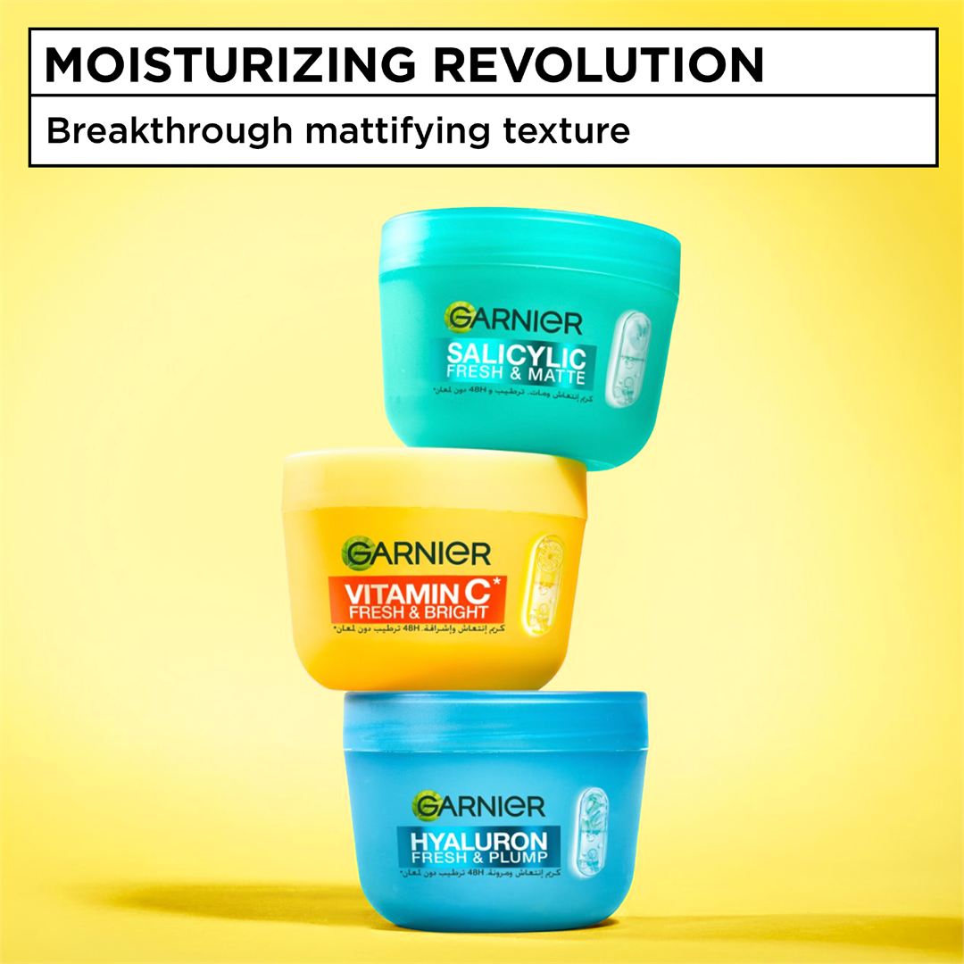 Three Garnier skincare jars stacked vertically on a yellow background with a white text banner at the top reading 'MOISTURIZING REVOLUTION' and 'Breakthrough mattifying texture.' The tubs are teal, yellow, and blue, labeled Salicylic Acid Fresh & Matte, Vitamin C Fresh & Bright, and Hyaluronic Acid Fresh & Plump.