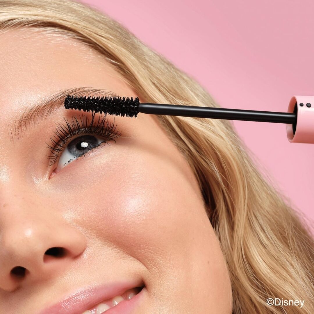 Person applying Essence alice in wonderland mascara to their eyelashes with a pink background